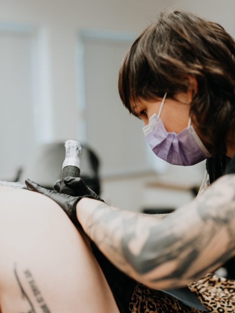 Tatowierer tätowiert einen Arm, trägt Handschuhe und eine Gesichtmaske.